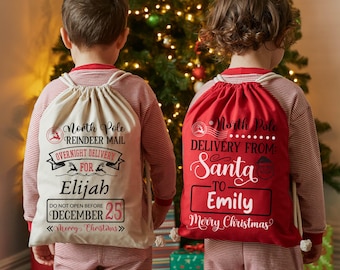 May include: Two drawstring Christmas sacks, one beige and one red, with festive text. The beige sack reads "North Pole Reindeer Mail" and "Overnight Delivery for Elijah." The red sack says "Delivery from Santa to Emily." Both say "Merry Christmas."