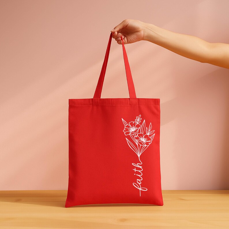 A hand holding a red tote bag with a floral design and the word "faith" written on it.