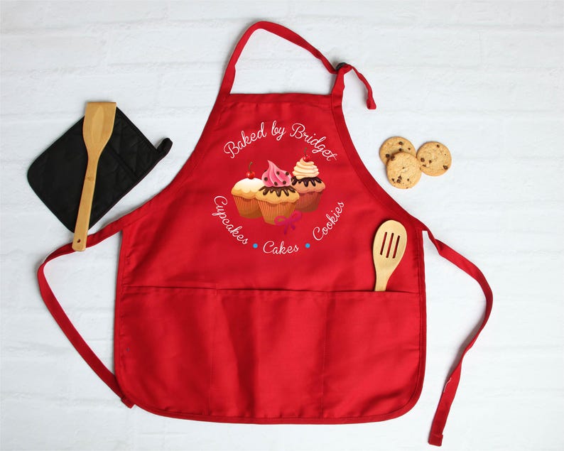 May include: A red apron with a pocket, wooden spoon, and oven mitt. The apron features the text "Baked by Bridget" and images of cupcakes. Chocolate chip cookies are on the right.