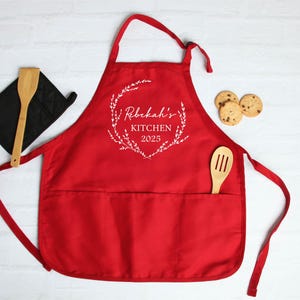 K&ouml;nnte beinhalten: Rote K&uuml;chensch&uuml;rze mit einer gro&szlig;en Tasche und verstellbaren Tr&auml;gern. Die Sch&uuml;rze hat ein wei&szlig;es Design mit der Aufschrift "Rebekah's Kitchen 2025" mit einem Kranz aus Bl&auml;ttern um den Text.