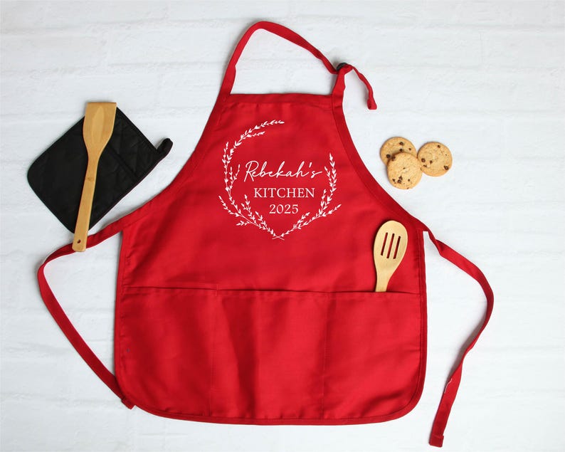 K&ouml;nnte beinhalten: Rote K&uuml;chensch&uuml;rze mit einer gro&szlig;en Tasche und verstellbaren Tr&auml;gern. Die Sch&uuml;rze hat ein wei&szlig;es Design mit der Aufschrift "Rebekah's Kitchen 2025" mit einem Kranz aus Bl&auml;ttern um den Text.