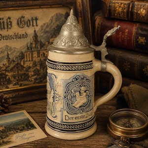 May include: A cream-colored ceramic beer stein with a detailed pewter lid and handle. The stein features blue embossed relief designs, including a cherub and the German text "Der erste Zug" on the front. The body has decorative borders.