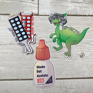 Może przedstawiać: Trzy naklejki przedstawiające dinozaura w okularach z napisem "PHO-RAPTOR", butelkę kropli do oczu z napisem "Shake Dat Acetate 1%" oraz dwie linijki z napisem "With the Rule" i "Against the Rule".