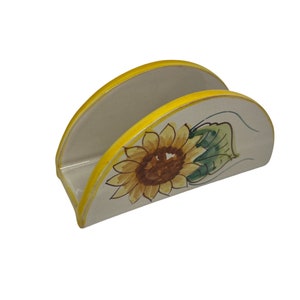 May include: Ceramic napkin holder with a sunflower design. The holder is white with a yellow rim and a painted sunflower with green leaves.
