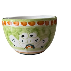 Könnte beinhalten: Eine kleine, handbemalte Keramikschale mit einem skurrilen Froschdesign. Die Schale hat eine grüne Außenseite mit einem sich wiederholenden Muster aus orangefarbenen und braunen Punkten am Rand. Der Frosch hat weiße Augen und einen lächelnden Mund.