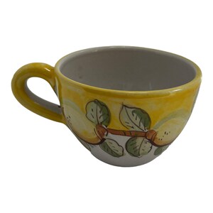 May include: A yellow ceramic teacup with a hand-painted design of lemons and leaves. The cup has a white interior and a rounded shape.
