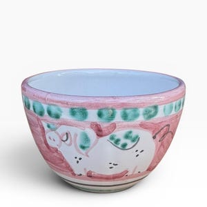 May include: A ceramic bowl with a hand-painted pig design. The bowl features a light blue interior, a pink rim with green polka dots, and a pink and white exterior with a pig illustration. The bowl is a decorative item.