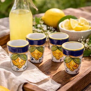 May include: Four small ceramic shot glasses with a yellow lemon design, blue rim, and white base sit on a wooden tray. A bottle of yellow liquid and a bowl of lemon slices are also present, suggesting a refreshing beverage.