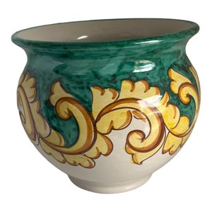 Puede incluir: Un macetero de cerámica blanca con un diseño floral verde y amarillo. El macetero tiene un cuerpo redondeado y un borde ancho.