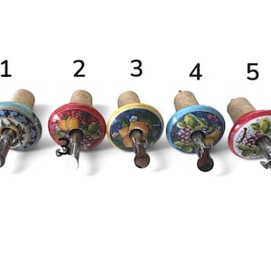 Può includere: Cinque tappi per bottiglie disposti in fila, numerati da uno a cinque. Ogni tappo presenta una parte superiore in ceramica rotonda dipinta a mano con motivi floreali e di frutta colorati, un beccuccio in metallo e una base in sughero.