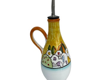 May include: Yellow ceramic oil and vinegar bottle with a hand-painted design of a small town with white buildings, green trees, and orange accents. The bottle has a silver spout and a cork stopper.