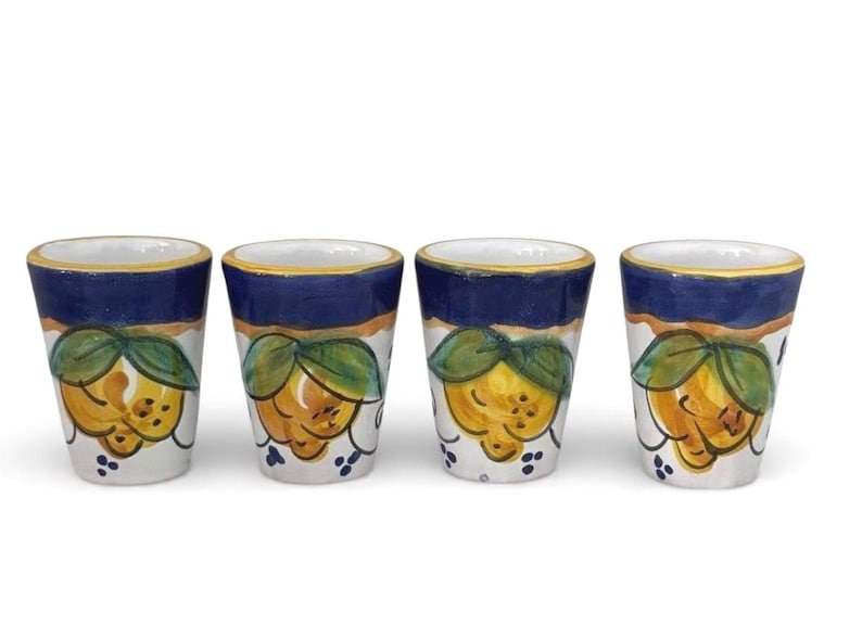 May include: Set of four ceramic shot glasses. Each glass features a white base with a hand-painted design of yellow lemons and green leaves. The top rim is navy blue with a gold accent.