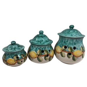 May include: Set of three ceramic canisters with lids, featuring a green and white design with yellow lemons and green leaves. The canisters have holes in the sides for ventilation.