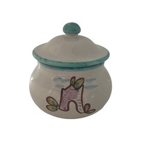 Op de afbeelding: Witte suikerpot van keramiek met een deksel. De pot is handgeschilderd met een ontwerp van een paars huis met groene bomen en blauwe wolken.