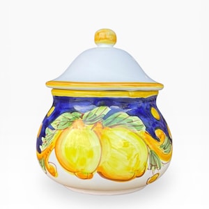 Op de afbeelding: Een keramische pot met deksel, versierd met een handgeschilderd citroen- en bladerenmotief. De pot heeft een witte basis, een blauwe body met gele accenten en een wit deksel met een gele knop. Italiaans aardewerk.
