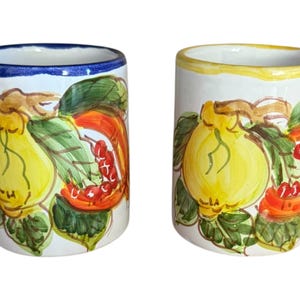 May include: Two white ceramic tumblers with hand-painted fruit designs. One has a blue rim, the other a yellow rim. The artwork features yellow lemons, green leaves, and red berries. The tumblers are likely for beverages.