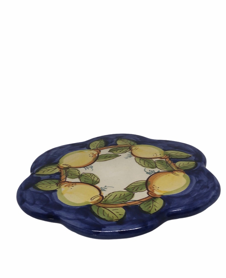 May include: A blue and white ceramic trivet with a flower-shaped design. The trivet is decorated with a wreath of lemons and green leaves.