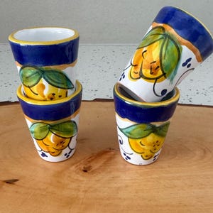 May include: Four ceramic shot glasses with a hand-painted lemon design. The glasses have a white base, blue rim, and yellow and green lemon illustrations. The glasses are stacked and arranged on a wooden surface.