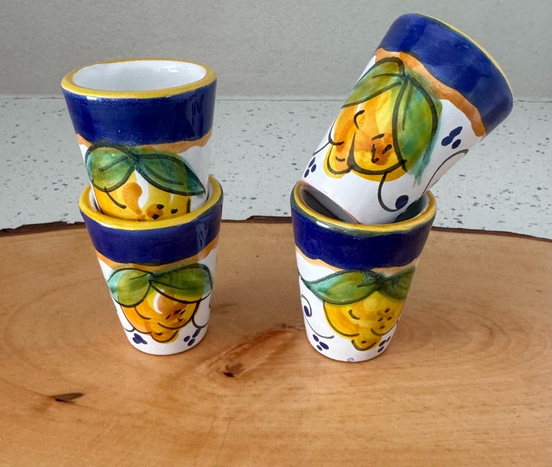 May include: Four ceramic shot glasses with a hand-painted lemon design. The glasses have a white base, blue rim, and yellow and green lemon illustrations. The glasses are stacked and arranged on a wooden surface.