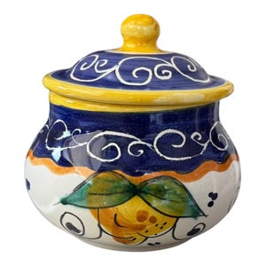 May include: A ceramic jar with a lid, featuring a hand-painted design. The jar has a white base with a yellow and orange band, a blue body, and a yellow lid. The lid is adorned with white swirls, and the jar depicts a lemon with green leaves.