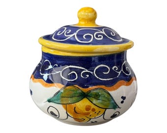 Peut inclure: Pot en céramique peint à la main avec couvercle. Le pot a une base blanche avec une bande jaune et orange, un corps bleu et un couvercle jaune. Le couvercle est orné de volutes blanches et le pot représente un citron avec des feuilles vertes.