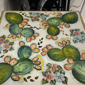 May include: A square table top with a white background and a painted design of green prickly pear cactus plants, red and yellow fruit, and pink and blue flowers. The table top is surrounded by a yellow rope border.