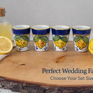 May include: Four ceramic shot glasses with a blue rim and a lemon design, arranged on a wooden board with a lemon and a bottle of liquid. The glasses have a white base with yellow lemons and green leaves.