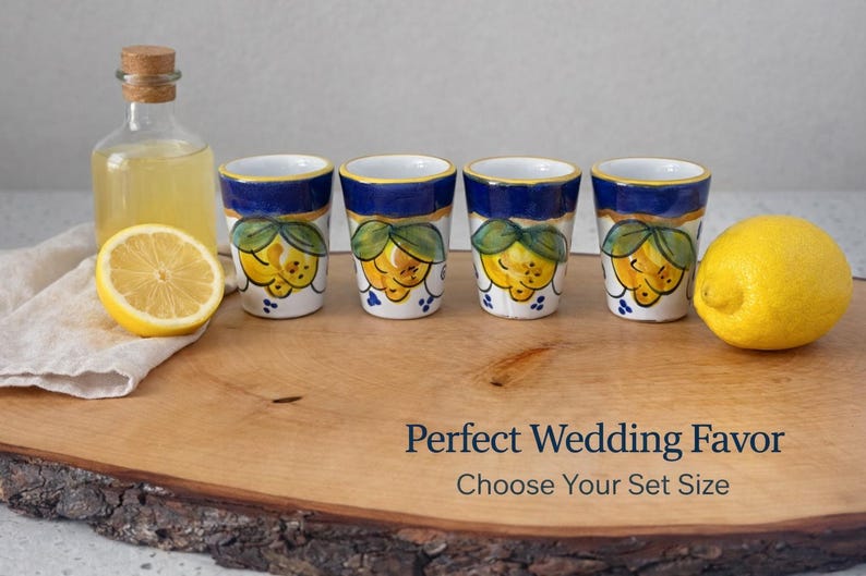 May include: Four ceramic shot glasses with a blue rim and a lemon design, arranged on a wooden board with a lemon and a bottle of liquid. The glasses have a white base with yellow lemons and green leaves.