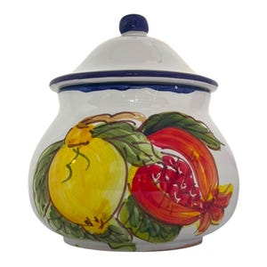 May include: A white ceramic jar with a lid, adorned with a hand-painted design of a yellow lemon and a red pomegranate with green leaves. The jar has a blue rim and a blue knob on the lid. A decorative kitchen storage container.
