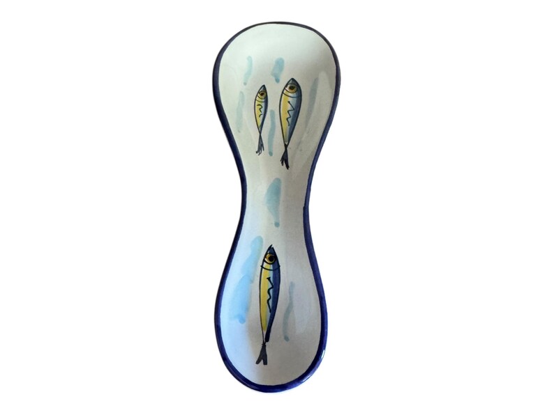 May include: A ceramic spoon rest with a white background, blue trim, and a whimsical fish design. The spoon rest features three yellow and blue fish illustrations and light blue water-like accents.