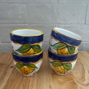 May include: A set of four ceramic bowls featuring a hand-painted design of yellow lemons with green leaves. Each bowl has a wide dark blue band around the rim and a white interior. The bowls are arranged in two stacks of two on a wooden surface.
