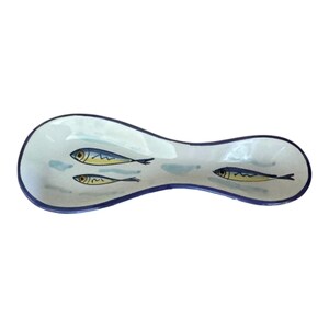 May include: A white ceramic spoon rest with a blue rim and three fish illustrations. The fish are blue and yellow with black details. The spoon rest is approximately 10 inches long.