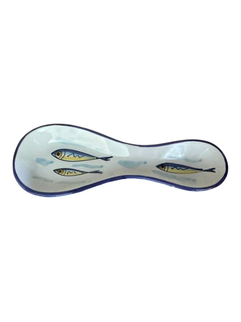 May include: A white ceramic spoon rest with a blue rim and three fish illustrations. The fish are blue and yellow with black details. The spoon rest is approximately 10 inches long.