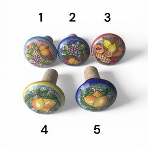 Puede incluir: Cinco tapones de botella decorativos con bases de corcho. Cada tapón presenta una parte superior de cerámica pintada a mano con un diseño de fruta diferente, que incluye uvas, limones y melocotones. Los tapones tienen una variedad de colores, incluyendo azul, amarillo y rojo.