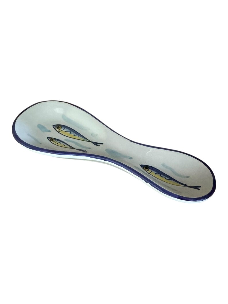 May include: A white ceramic spoon rest with a blue rim and three stylized fish illustrations. The fish are yellow and blue, and the spoon rest has a curved, elongated shape.