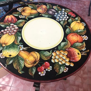 Op de afbeelding: Een ronde zwarte tafelblad met een geschilderd ontwerp van fruit en bloemen. Het tafelblad is omgeven door een witte rand. Het tafelblad is gemaakt van keramiek.