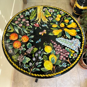 May include: A round black table top with a yellow rope border. The table top is painted with a colorful floral design featuring lemons, grapes, pears, and other fruits.