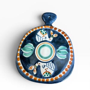 May include: A hand-painted, ceramic serving board with a dark blue background and a decorative design. The design includes white bird-like figures, green and orange accents, and a ring of orange dots around the edge. The board has a handle.