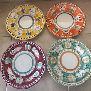May include: Four decorative ceramic plates with scalloped edges. Each plate features a unique design with fish, crabs, rabbits, and floral motifs in yellow, orange, burgundy, and green. The plates have a white center and a blue or orange border.