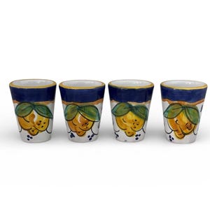 May include: Four ceramic shot glasses. Each glass features a hand-painted design of a yellow lemon with green leaves on a white background. The top of each glass has a blue band with a gold rim.