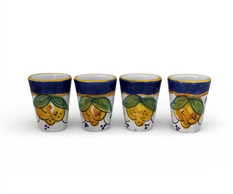 May include: Four ceramic shot glasses. Each glass features a hand-painted design of a yellow lemon with green leaves on a white background. The top of each glass has a blue band with a gold rim.