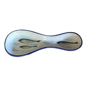 May include: A ceramic spoon rest with a light blue and white background and a dark blue rim. The spoon rest features three yellow and black fish illustrations.