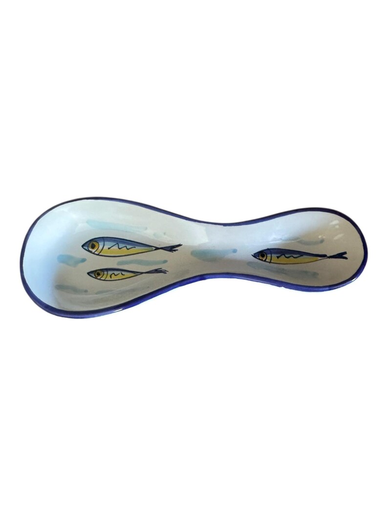May include: A ceramic spoon rest with a light blue and white background and a dark blue rim. The spoon rest features three yellow and black fish illustrations.