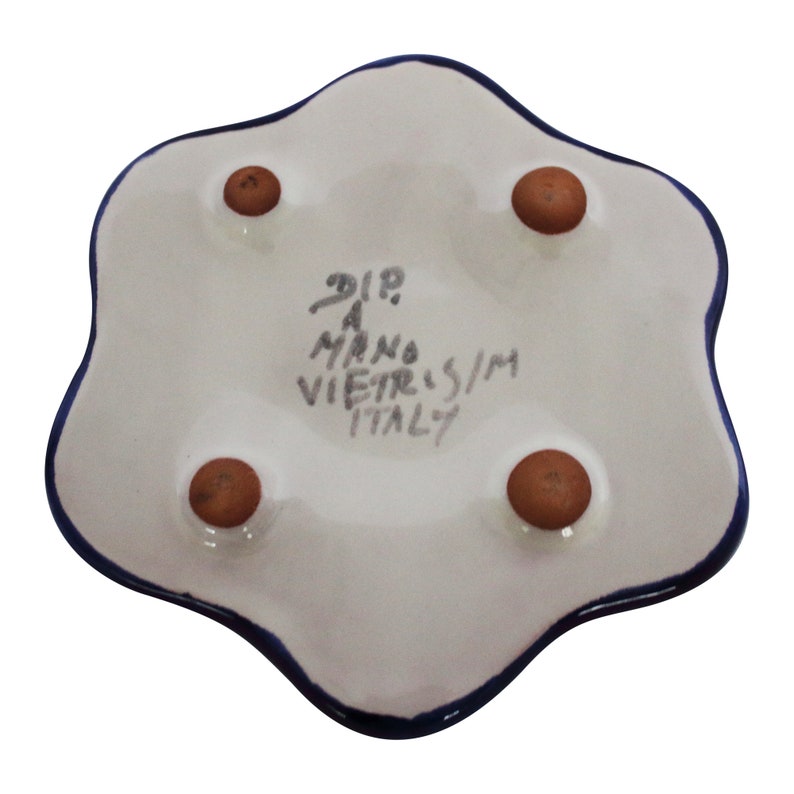 May include: A white ceramic dish with a blue rim and four brown feet. The bottom of the dish has the text "DIP A MANO VIETRIS/M ITALY" written in black.