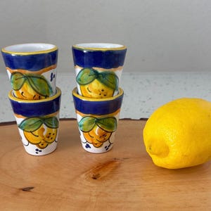 May include: Four small, hand-painted ceramic shot glasses with a lemon design. The glasses have a white base, blue rim, and a yellow and green lemon motif. A fresh yellow lemon sits next to the glasses on a wooden surface.