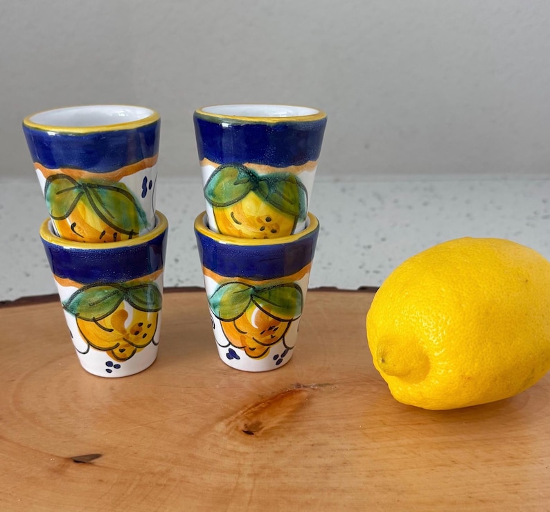 May include: Four small, hand-painted ceramic shot glasses with a lemon design. The glasses have a white base, blue rim, and a yellow and green lemon motif. A fresh yellow lemon sits next to the glasses on a wooden surface.