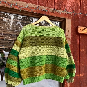 May include: A green, brown and cream striped crocheted sweater with long sleeves and a relaxed fit. The sweater is hanging on a wooden hanger.