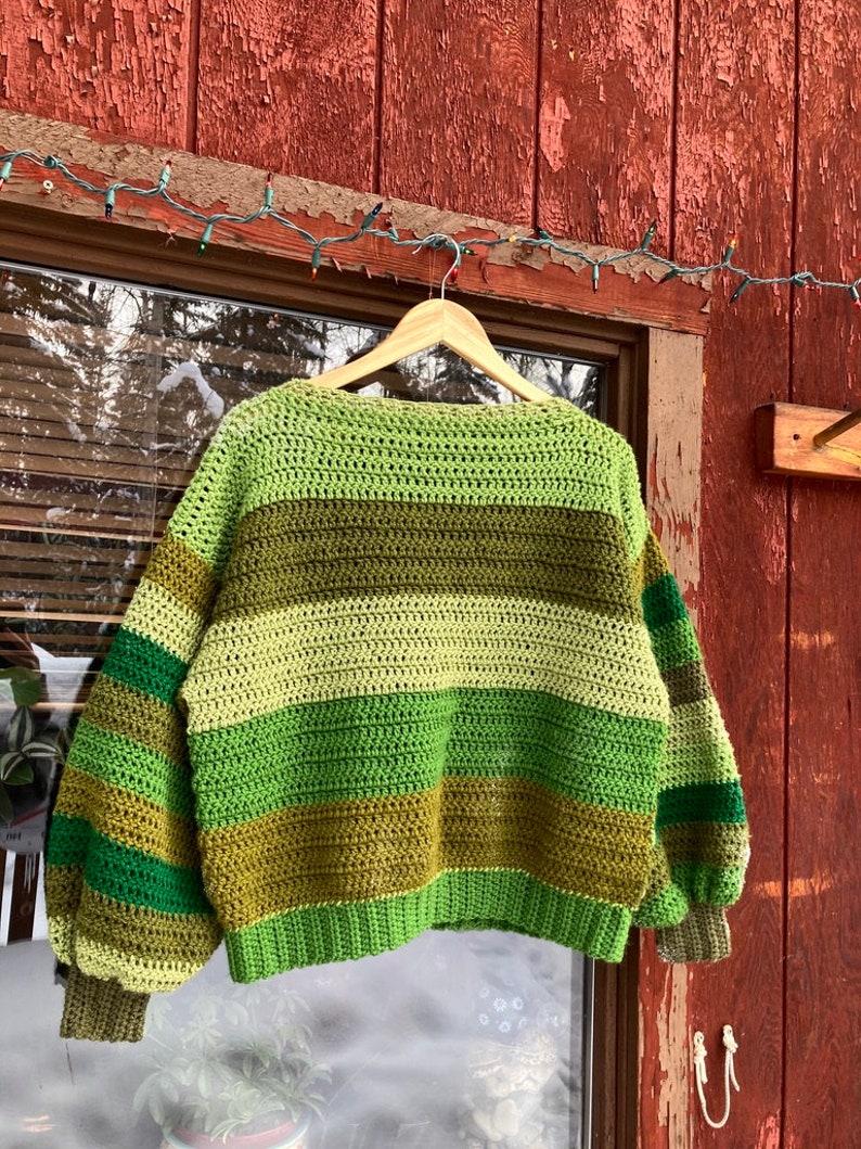 May include: A green, brown and cream striped crocheted sweater with long sleeves and a relaxed fit. The sweater is hanging on a wooden hanger.