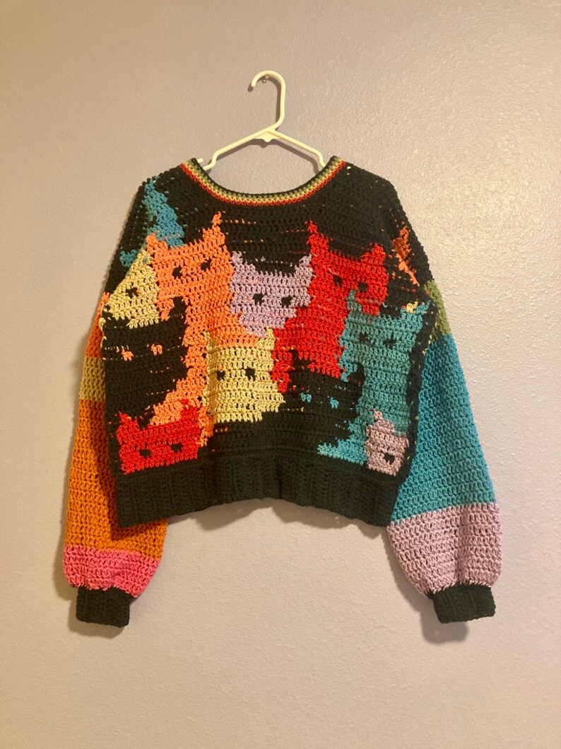 May include: A colourful crocheted sweater with a cat pattern. The sweater is black with multi-coloured cat faces and has long sleeves with a crocheted cuff.