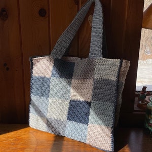 May include: A crocheted tote bag with a checkered pattern in shades of blue, grey, and white. The bag has two long handles.